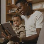 A father reads a book to his baby while sitting on a couch, creating a warm family moment.