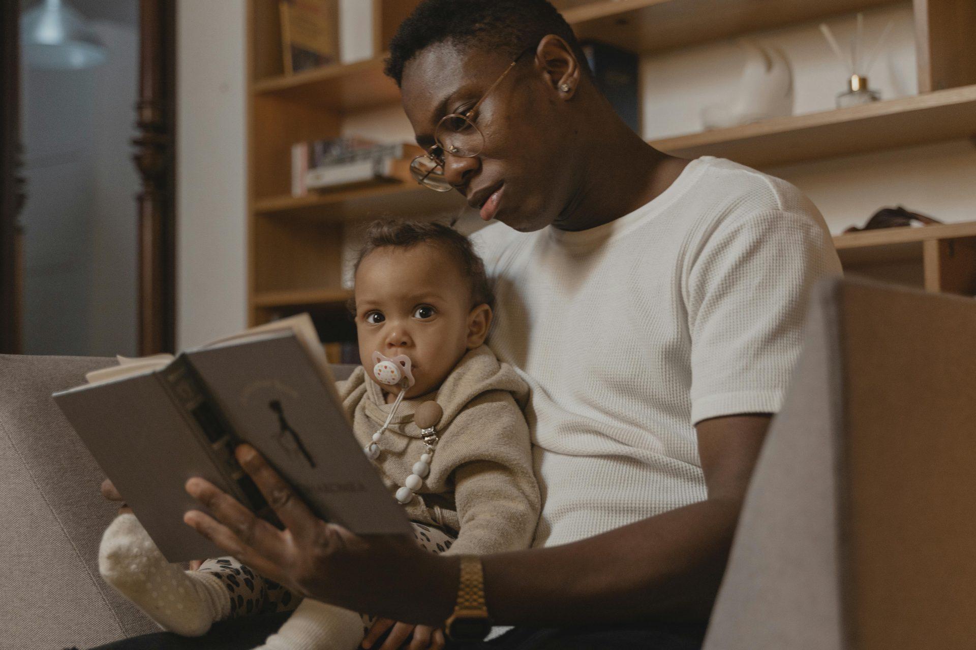 A father reads a book to his baby while sitting on a couch, creating a warm family moment.